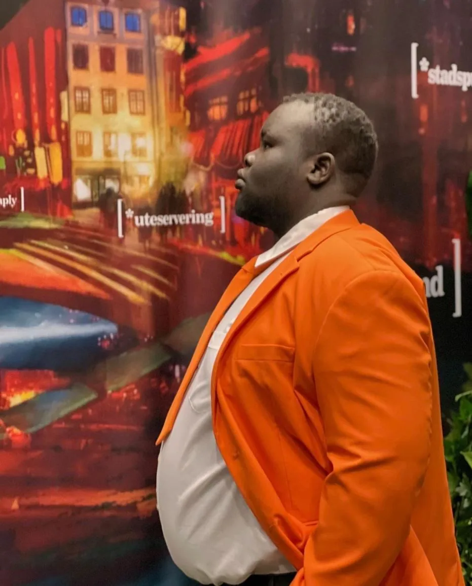 Megic Mike i orange kavaj och vit skjorta i profil framför en färgstark stadsmålning